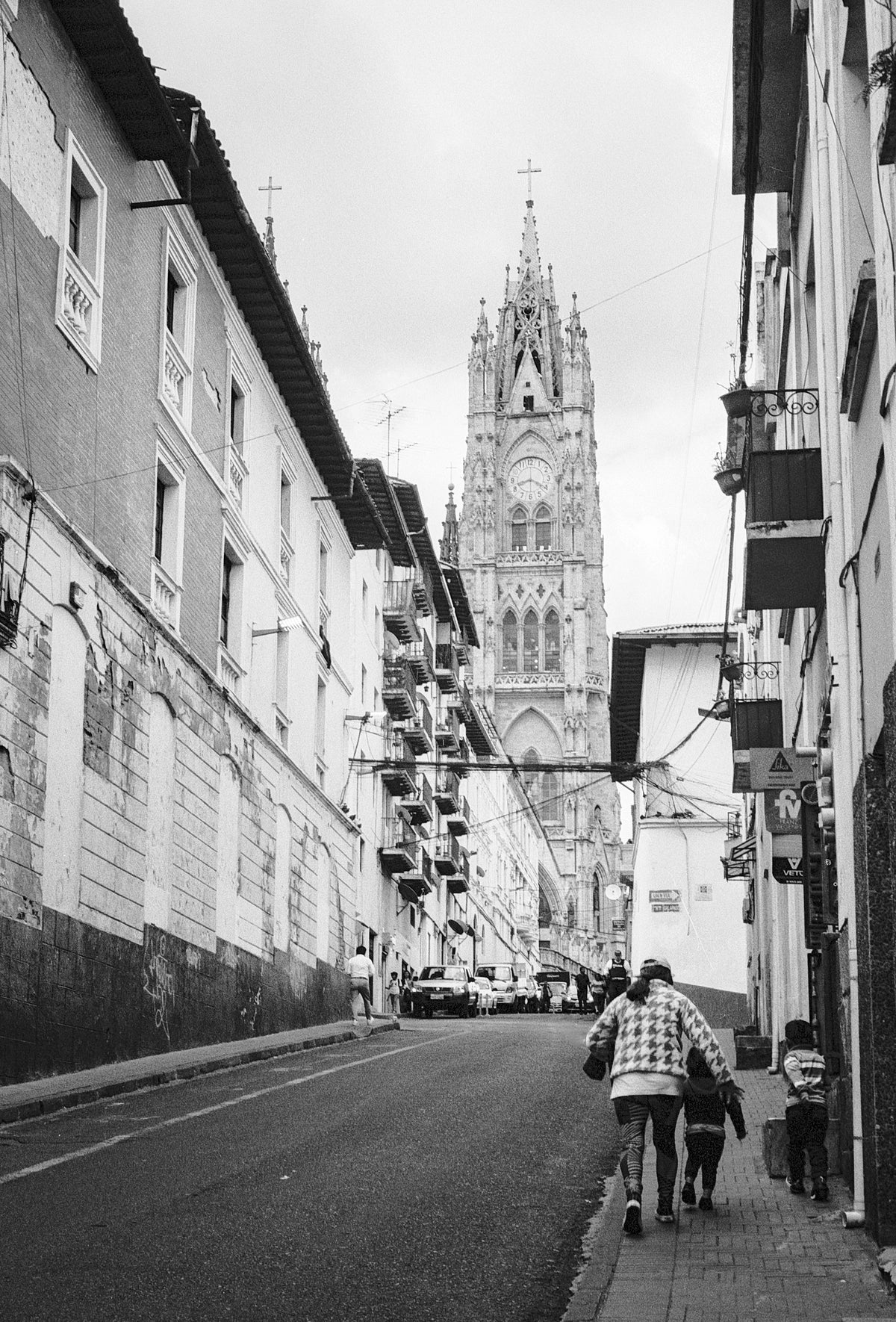 Black and white street photograph of a narrow urban road leading toward a tall cathedral spire, with buildings lining both sides and a few pedestrians walking in the foreground.